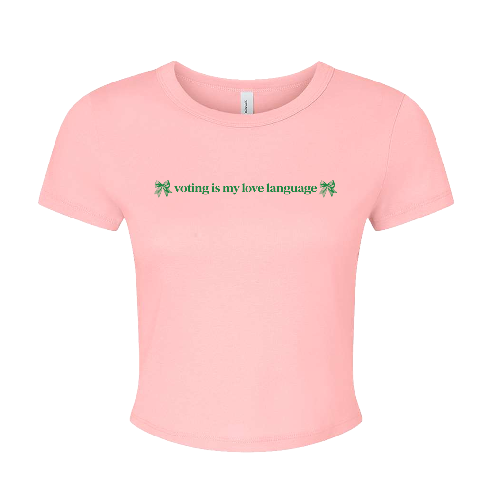A light pink short-sleeve crop top t-shirt with the green text 'voting is my love language' and small green bow graphics on the chest, displayed on a solid white background.