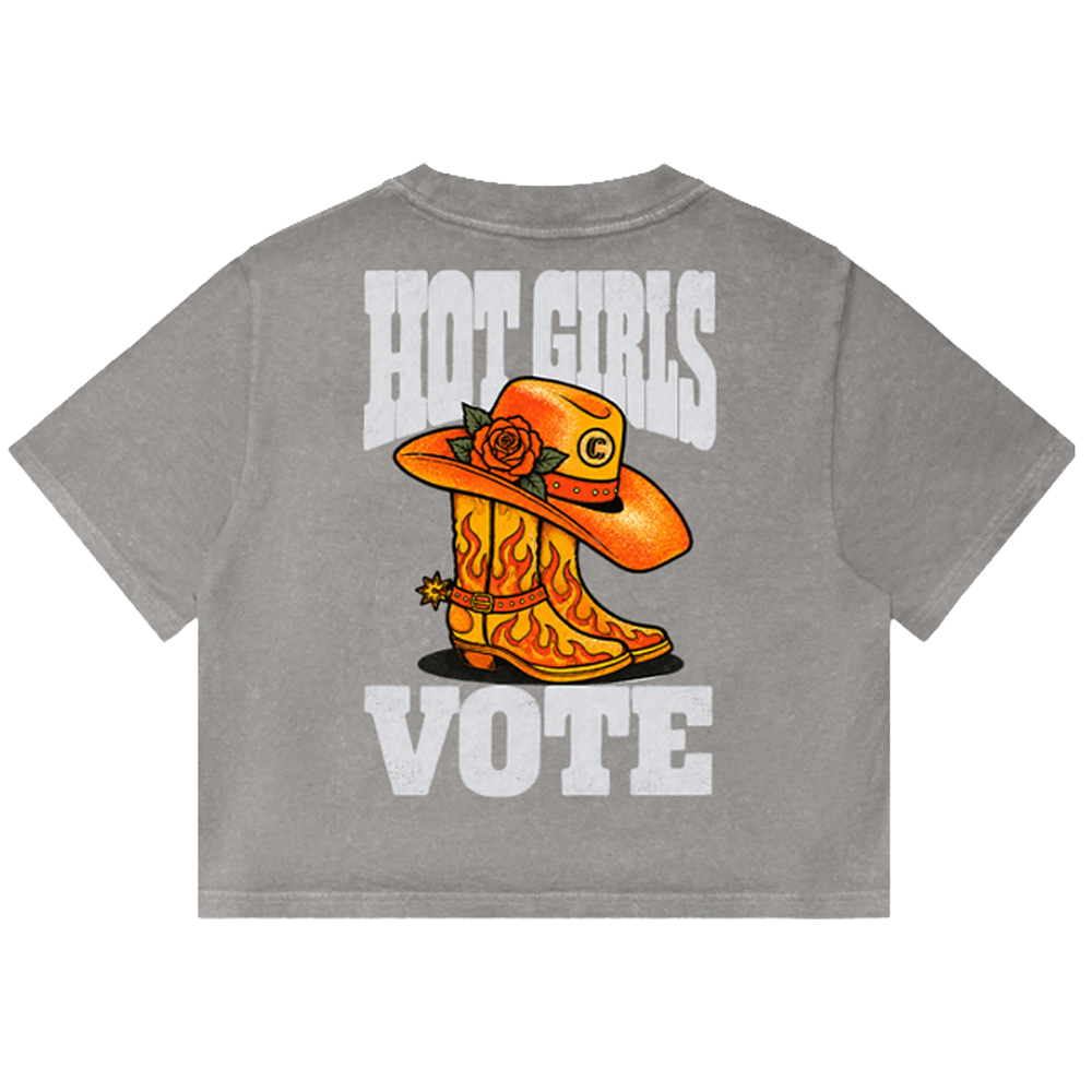 A gray short-sleeve t-shirt shown from the back, featuring a graphic print of an orange cowboy boot with flames, a matching orange cowboy hat with a rose, and the words 'HOT GIRLS VOTE' in distressed white lettering.