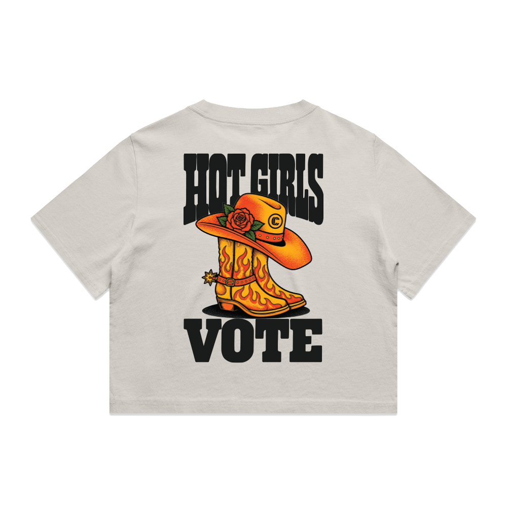 A light beige short-sleeve t-shirt shown from the back, featuring a graphic print of two orange cowboy boots with flames, a cowboy hat, a rose, and the text 'HOT GIRLS VOTE' in black.