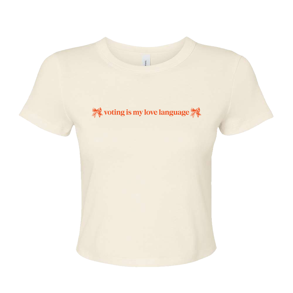 An off-white cropped short-sleeve t-shirt with orange text reading 'voting is my love language' flanked by two orange bow illustrations, displayed on a solid white background.